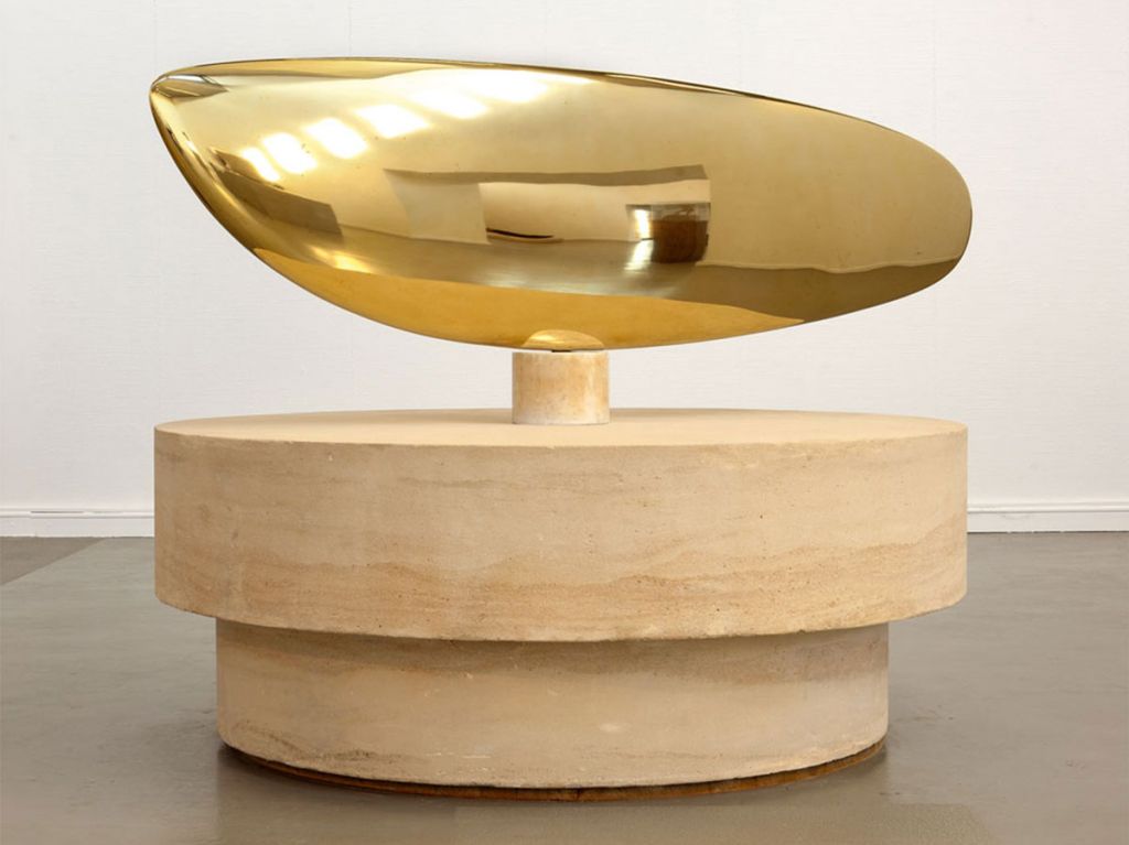Constantin Brâncuşi (*1876 - 1957), "Le grand poisson", 1930, Kunsthalle Mannheim / VG Bild-Kunst, Bonn 2023 Zu sehen ist eine goldglänzende Plastik aus polierter Bronze. Die geschwungene Form, die an den Körpers eines Fischs erinnert, sitzt auf einem großen hellbraunen Sockel aus Stein.