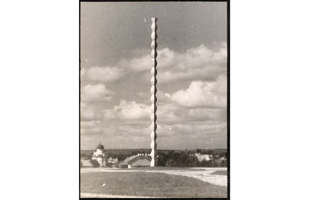 Die Schwarz-Weiß-Fotografie zeigt die Unendliche Säule auf einem begrünten Platz stehend. Im Hintergrund, weiter unten liegend, sind Gebäude zu erkennen.