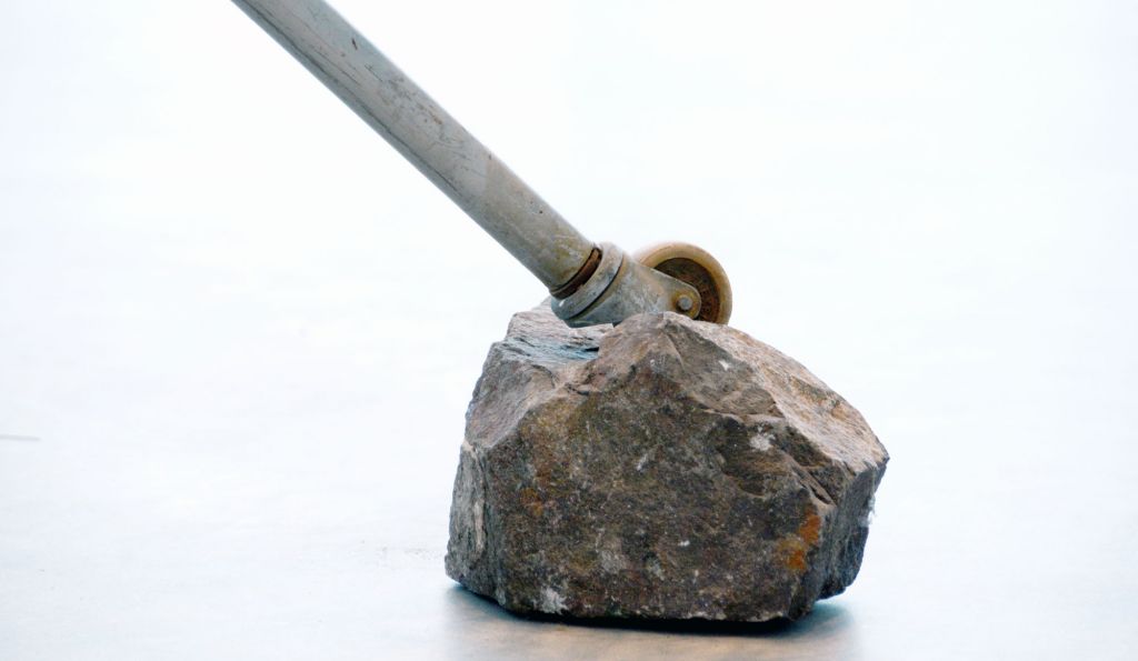 Rebecca Horn, Inferno, 1993, 11 Metallbetten, 4 Geigen, Motoren, Kunsthalle Mannheim © VG Bild-Kunst, Bonn 2023 (Foto: Kunsthalle Mannheim / Lukac Diehl) Die Detailaufnahme zeigt das Metallbein eines Bettes, das schräg auf einem grauen unbehauenem Stein steht. Das Metallbein mündet in einem kleinen Rad.