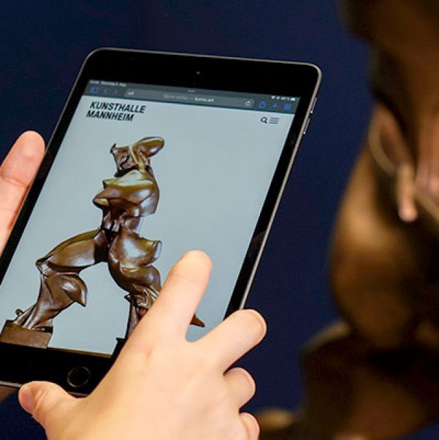 Das Foto zeigt Hände, die ein Tablet mit dem Modul zu Umberto Boccioni vor der Plastik Einzigartige Formen der Kontinuität im Raum halten.
