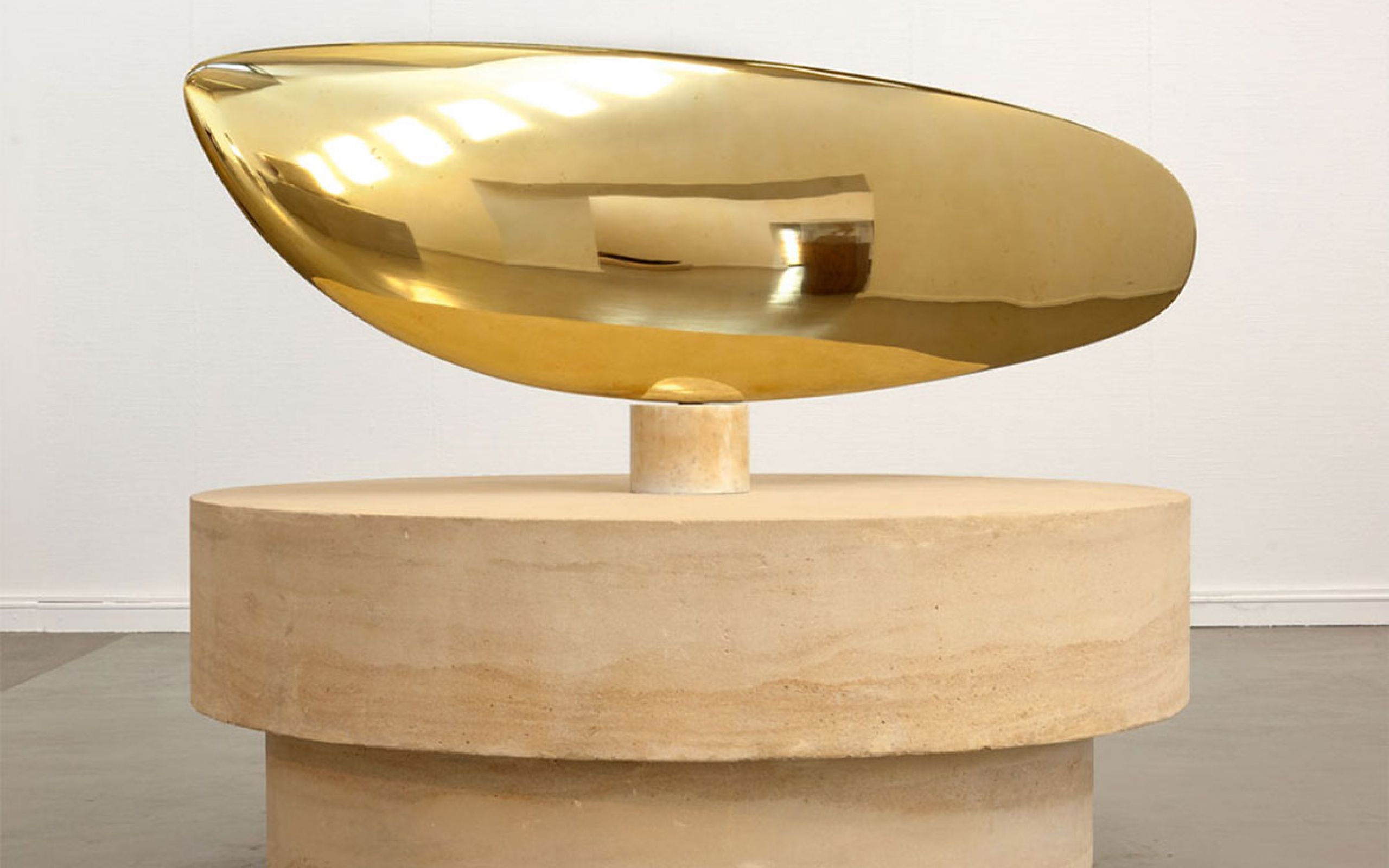 Zu sehen ist eine goldglänzende Plastik aus polierter Bronze. Die geschwungene Form, die an den Körpers eines Fischs erinnert, sitzt auf einem großen hellbraunen Sockel aus Stein.